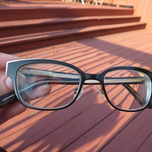 May include: Black and silver-framed eyeglasses with clear lenses. The glasses have a rectangular shape with a silver accent on the top of the frame. The temples are dark with a subtle design. The glasses are held against a wooden background.