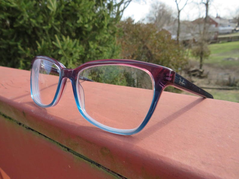 Puede incluir: Gafas con montura degradada, pasando de rojo oscuro a azul claro. Las patillas son negras y las lentes transparentes. Las gafas est&aacute;n apoyadas en una superficie.