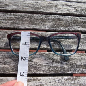 Puede incluir: Gafas con montura degradada, que transiciona del burdeos al azul. Las gafas se miden con una regla, mostrando una altura de aproximadamente 7 cm. Las monturas tienen forma rectangular.