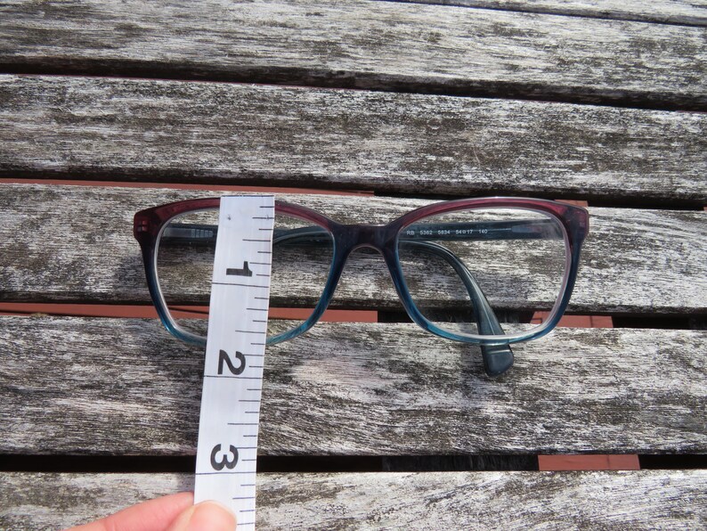 Puede incluir: Gafas con montura degradada, que transiciona del burdeos al azul. Las gafas se miden con una regla, mostrando una altura de aproximadamente 7 cm. Las monturas tienen forma rectangular.