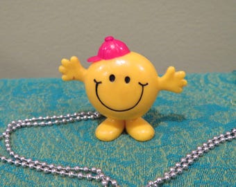 May include: A yellow figurine with a smiling face and a pink cap. The figurine has outstretched arms and small feet. A silver ball chain necklace is in the foreground. The figurine is on a teal patterned surface.