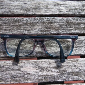 Puede incluir: Un par de gafas Ray-Ban con montura azul oscuro y morada, sobre una superficie de madera envejecida. Las lentes son transparentes y las patillas se curvan hacia abajo. El logotipo de Ray-Ban es visible en la esquina superior izquierda de la montura.