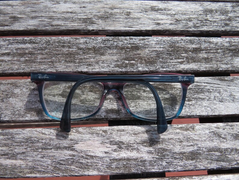 Puede incluir: Un par de gafas Ray-Ban con montura azul oscuro y morada, sobre una superficie de madera envejecida. Las lentes son transparentes y las patillas se curvan hacia abajo. El logotipo de Ray-Ban es visible en la esquina superior izquierda de la montura.
