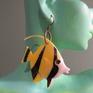 May include: A pair of dangle earrings featuring a yellow and black fish design. The fish is made of plastic and has a white belly. The earrings are hanging from a silver hook.