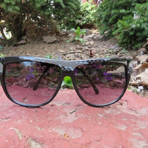 May include: Black sunglasses with a geometric pattern on the frame. The lenses are dark and reflect the surrounding environment.