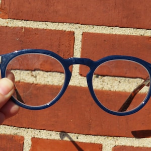 May include: Round, navy blue eyeglasses with clear lenses are held against a red brick wall. The frames have a subtle, modern design. The temples are a tortoiseshell pattern. The glasses are in focus, with the brick background slightly blurred.