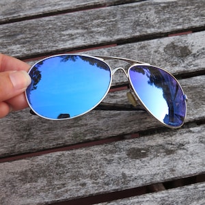 May include: Aviator sunglasses featuring a thin silver metal frame and reflective blue lenses. The lenses show a clear reflection of trees and the sky. The bridge of the frame has a double bar design, and the nose pads are a light beige color.