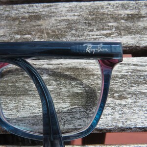 Puede incluir: Primer plano de gafas Ray-Ban. Las monturas son un degradado de azul y rojo, con el logotipo de Ray-Ban visible en la patilla. Las gafas descansan sobre una superficie de madera envejecida.