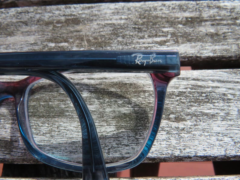 Puede incluir: Primer plano de gafas Ray-Ban. Las monturas son un degradado de azul y rojo, con el logotipo de Ray-Ban visible en la patilla. Las gafas descansan sobre una superficie de madera envejecida.