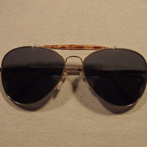 May include: Aviator sunglasses with dark lenses and a gold-colored metal frame. The top bar features a tortoiseshell pattern. The sunglasses have a classic design, suitable for various styles and occasions.