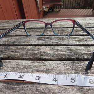 Puede incluir: Gafas con montura degradada, que va del rojo en la parte superior al azul en la inferior. Las patillas son de color azul oscuro. Debajo de las gafas hay una cinta m&eacute;trica que muestra pulgadas.