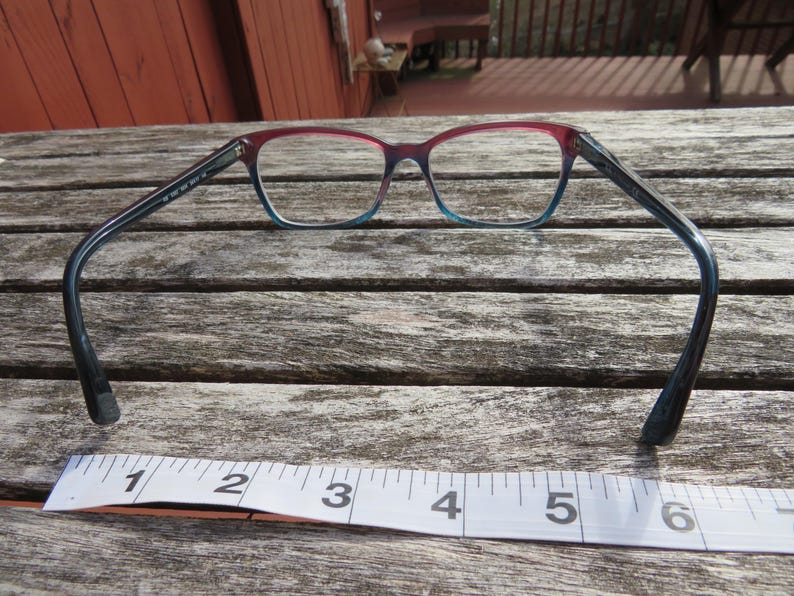 Puede incluir: Gafas con montura degradada, que va del rojo en la parte superior al azul en la inferior. Las patillas son de color azul oscuro. Debajo de las gafas hay una cinta m&eacute;trica que muestra pulgadas.