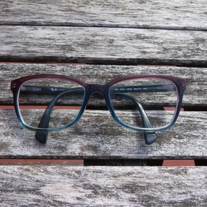 Puede incluir: Gafas rectangulares con montura degradada, que va del burdeos al azul. Las monturas tienen un dise&ntilde;o cl&aacute;sico, con el logotipo de Ray-Ban visible en la patilla. Las gafas est&aacute;n sobre una superficie de madera envejecida.