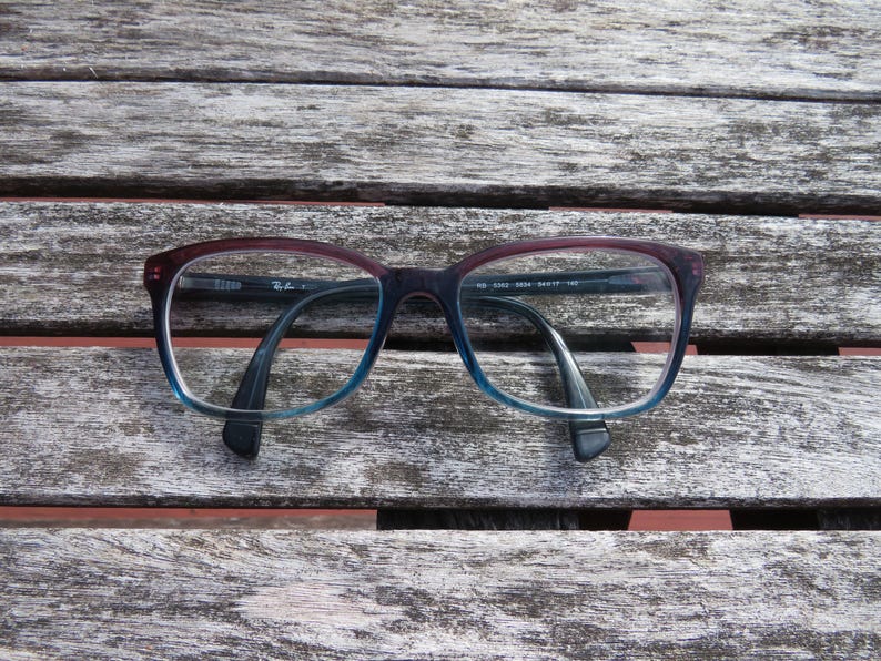 Puede incluir: Gafas rectangulares con montura degradada, que va del burdeos al azul. Las monturas tienen un dise&ntilde;o cl&aacute;sico, con el logotipo de Ray-Ban visible en la patilla. Las gafas est&aacute;n sobre una superficie de madera envejecida.