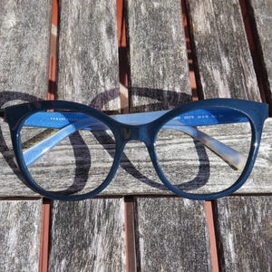May include: Blue cat-eye glasses with a blue frame and clear lenses. The glasses are sitting on a wooden surface.