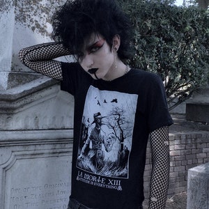May include: A person wearing a black t-shirt with a white graphic print of a figure wearing a gas mask and holding a skull. The t-shirt has the text "La Morte XIII" and "Attitude is Everything" printed on it. The person is also wearing black fishnet sleeves and blue jeans.