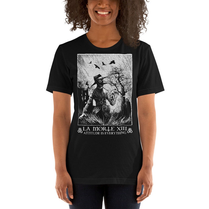 May include: Black t-shirt with a white graphic print of a person wearing a plague doctor mask and holding a skull. The text "LA MORTE XIII" and "ATTITUDE IS EVERYTHING" is printed below the graphic.