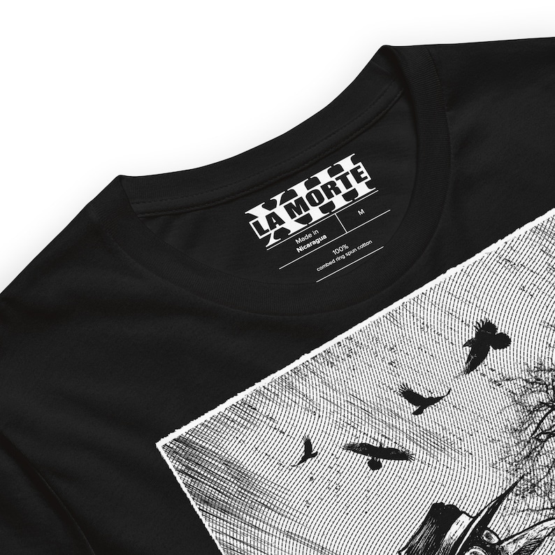 May include: Black t-shirt with a white graphic print of a person wearing a hat and crows flying in the sky. The shirt has a white tag that says "LA MORTE" and "Made in Nicaragua 100% combed ring spun cotton".