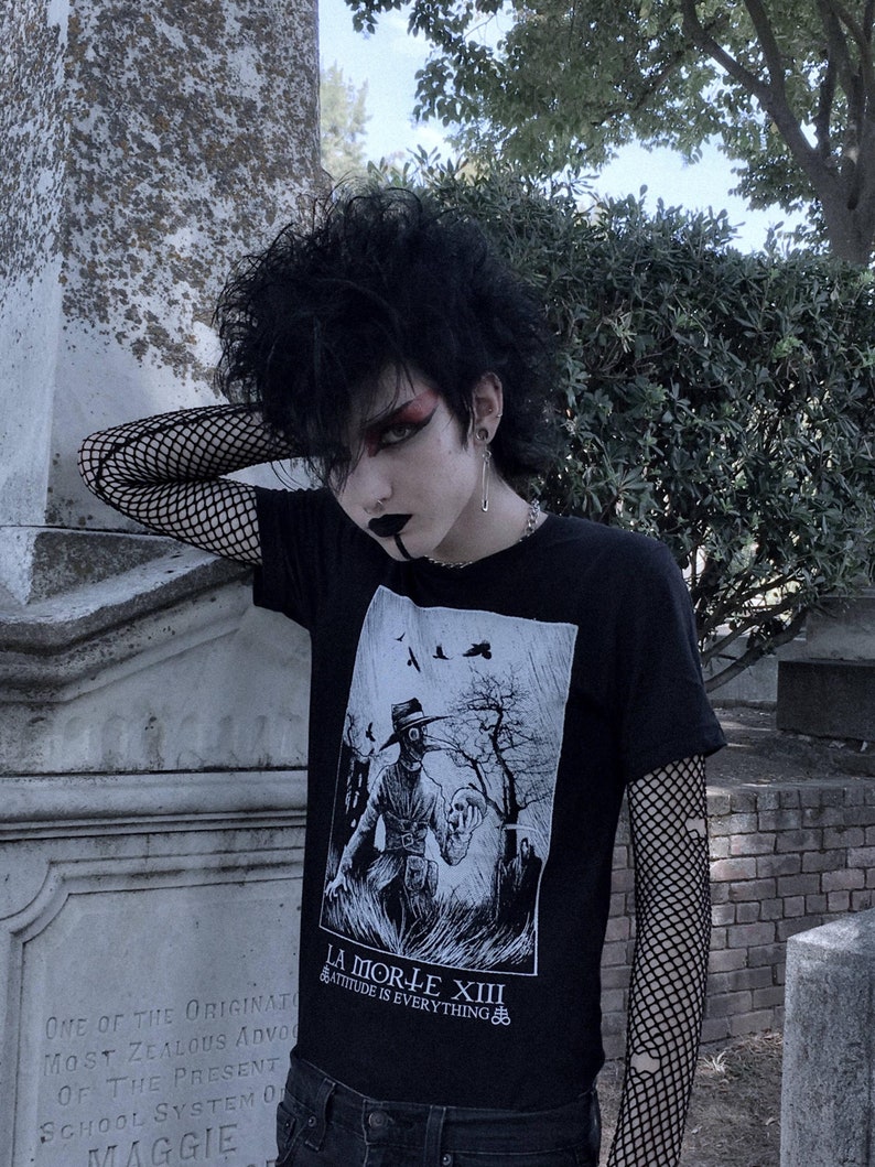 May include: A person wearing a black t-shirt with a white graphic print of a figure wearing a gas mask and holding a skull. The t-shirt has the text "La Morte XIII" and "Attitude is Everything" printed on it. The person is also wearing black fishnet sleeves and blue jeans.