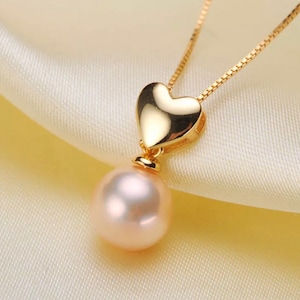 May include: A gold heart pendant necklace with a pink pearl drop. The necklace is on a white satin background.