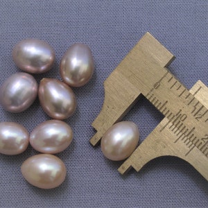 Puede incluir: Un grupo de siete perlas ovaladas, iridiscentes, de color rosa claro con una regla que mide una de las perlas a 1,8 cm.