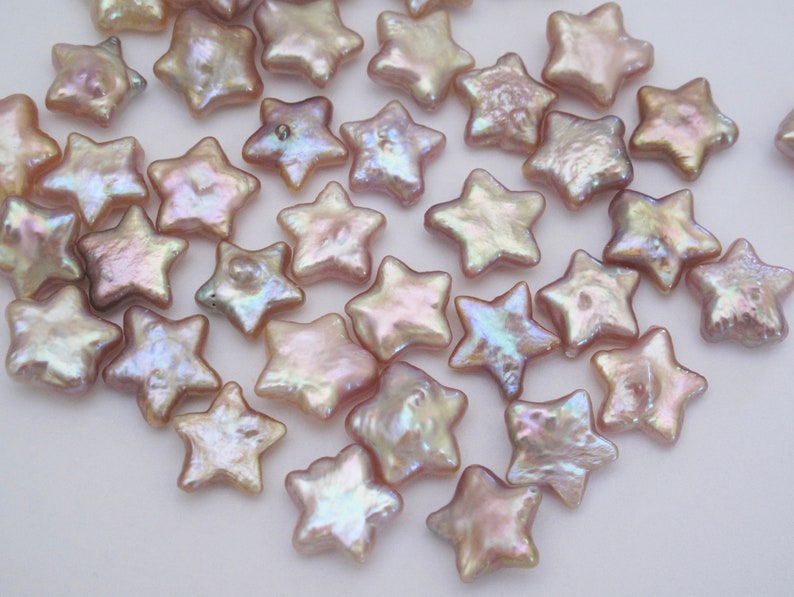 May include: A collection of iridescent pink star-shaped pearl beads. The beads are scattered on a white surface.