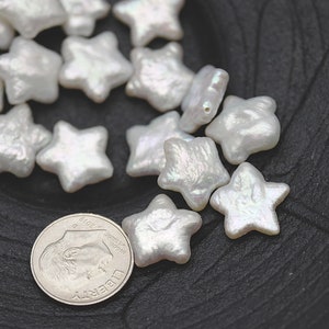 May include: A collection of white pearl star-shaped beads. The beads are iridescent and have a slightly irregular shape. The beads are scattered on a black surface.