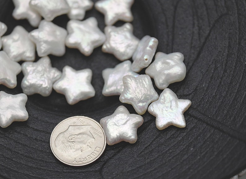 May include: A collection of white pearl star-shaped beads. The beads are iridescent and have a slightly irregular shape. The beads are scattered on a black surface.