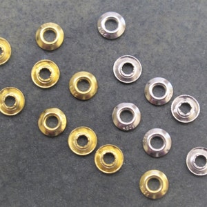 May include: Assortment of metal eyelets in gold and silver finishes. The eyelets are arranged in a diagonal pattern on a gray surface.