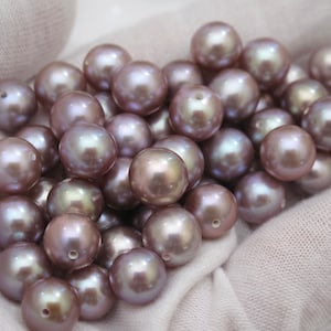 May include: A collection of lustrous, round pearls in shades of mauve and silver. Each pearl has a visible hole, suggesting they are intended for stringing into jewelry. The pearls are displayed on a soft, white fabric.