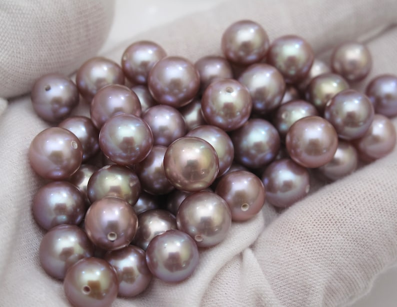 May include: A collection of lustrous, round pearls in shades of mauve and silver. Each pearl has a visible hole, suggesting they are intended for stringing into jewelry. The pearls are displayed on a soft, white fabric.