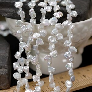May include: Strands of white, irregularly shaped freshwater pearls. The pearls are strung together, creating a necklace or bracelet. The pearls have a lustrous sheen and are displayed near a white ceramic cup and a ruler.