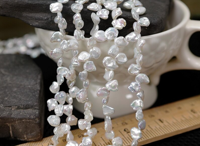 May include: Strands of white, irregularly shaped freshwater pearls. The pearls are strung together, creating a necklace or bracelet. The pearls have a lustrous sheen and are displayed near a white ceramic cup and a ruler.