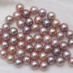 May include: A collection of round, iridescent pearls in shades of lavender, pink, and white. The pearls are scattered on a white surface.