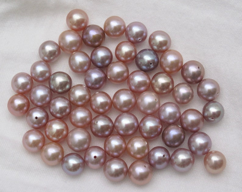 May include: A collection of round, iridescent pearls in shades of lavender, pink, and white. The pearls are scattered on a white surface.