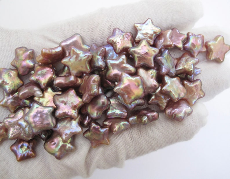 May include: A pile of iridescent pink and gold star-shaped pearl beads. The beads are arranged in a loose pile on a white surface.