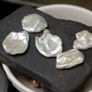 May include: Five lustrous, irregularly shaped, white baroque pearls rest on a dark gray, textured stone. The pearls have a pearlescent sheen, reflecting light. The stone sits inside a white bowl, suggesting a jewelry or craft theme.