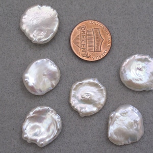 May include: Six white, round, irregular shaped, iridescent pearls, with one pearl slightly larger than the others, next to a US penny.