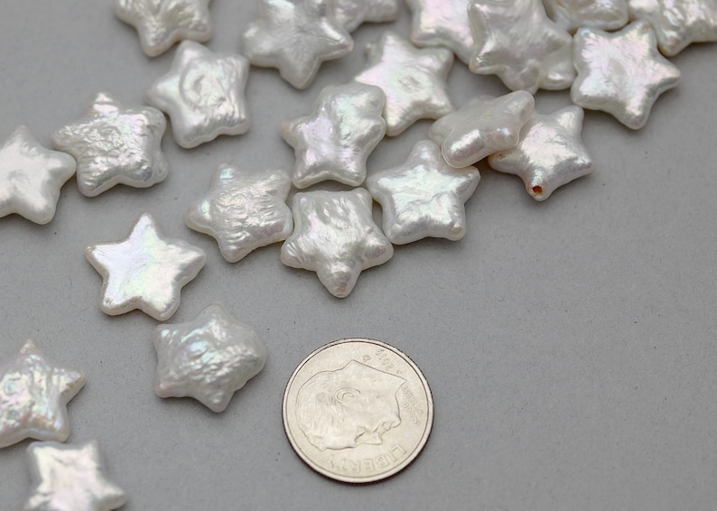 May include: A collection of white pearl star-shaped beads. The beads are iridescent and have a slightly textured surface. The beads are scattered on a gray surface next to a US quarter.