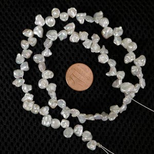 May include: Strand of irregular, off-white freshwater pearls. The pearls are strung on a thin, white thread. A U.S. one-cent coin is next to the pearls for size comparison. The pearls have a lustrous sheen and vary in shape and size.