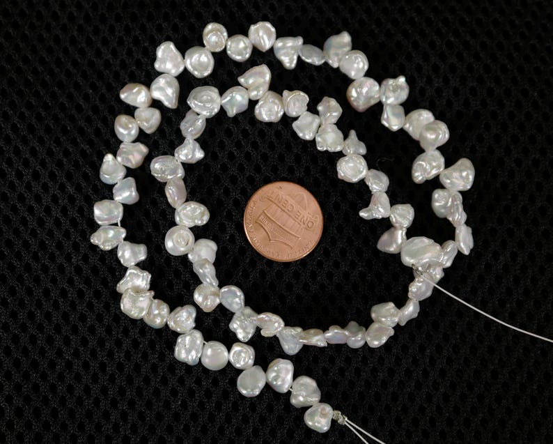 May include: Strand of irregular, off-white freshwater pearls. The pearls are strung on a thin, white thread. A U.S. one-cent coin is next to the pearls for size comparison. The pearls have a lustrous sheen and vary in shape and size.