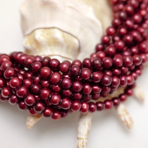 May include: A strand of deep red freshwater pearls, arranged in a circular pattern, with a white seashell in the background.