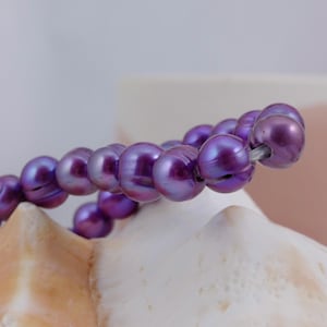 Puede incluir: Una hebra de perlas de agua dulce de color púrpura con un brillo iridiscente. Las perlas están enhebradas en un hilo transparente y están dispuestas en un patrón suelto y fluido.
