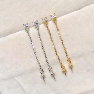 Puede incluir: Dos pares de cierres de pendientes con un pequeño cristal cuadrado transparente en la parte superior y una cadena dorada o plateada que se extiende hasta una tapa de perla.