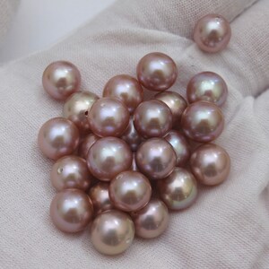 May include: A collection of lustrous, round, mauve-colored pearls. The pearls are scattered on a white surface, with a soft sheen and subtle color variations. These beads are ideal for jewelry making.