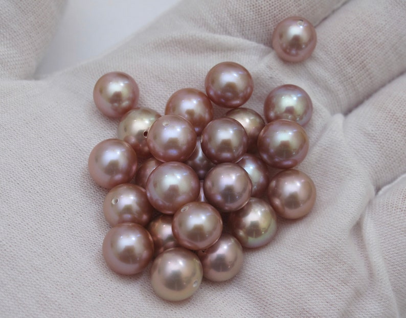 May include: A collection of lustrous, round, mauve-colored pearls. The pearls are scattered on a white surface, with a soft sheen and subtle color variations. These beads are ideal for jewelry making.