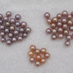 May include: A collection of lustrous, round pearls in shades of lavender, peach, and champagne. The pearls are arranged in three separate groups, each displaying a unique color variation and natural sheen. The pearls are on a white, textured surface.