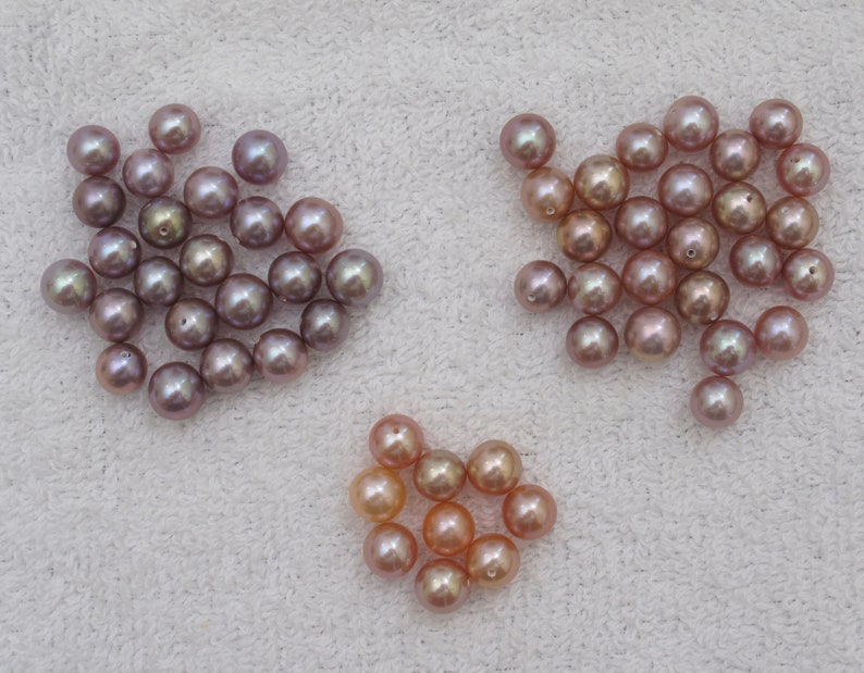 May include: A collection of lustrous, round pearls in shades of lavender, peach, and champagne. The pearls are arranged in three separate groups, each displaying a unique color variation and natural sheen. The pearls are on a white, textured surface.
