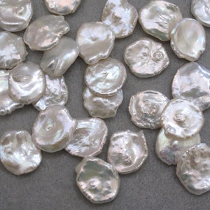 May include: A collection of white, iridescent, round, flat, irregular shaped pearls. The pearls are scattered on a gray surface.
