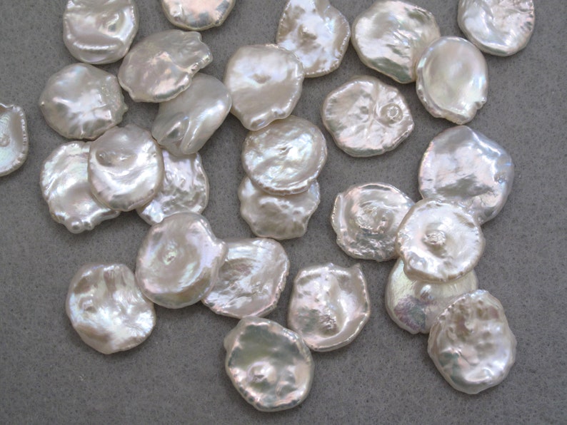 May include: A collection of white, iridescent, round, flat, irregular shaped pearls. The pearls are scattered on a gray surface.